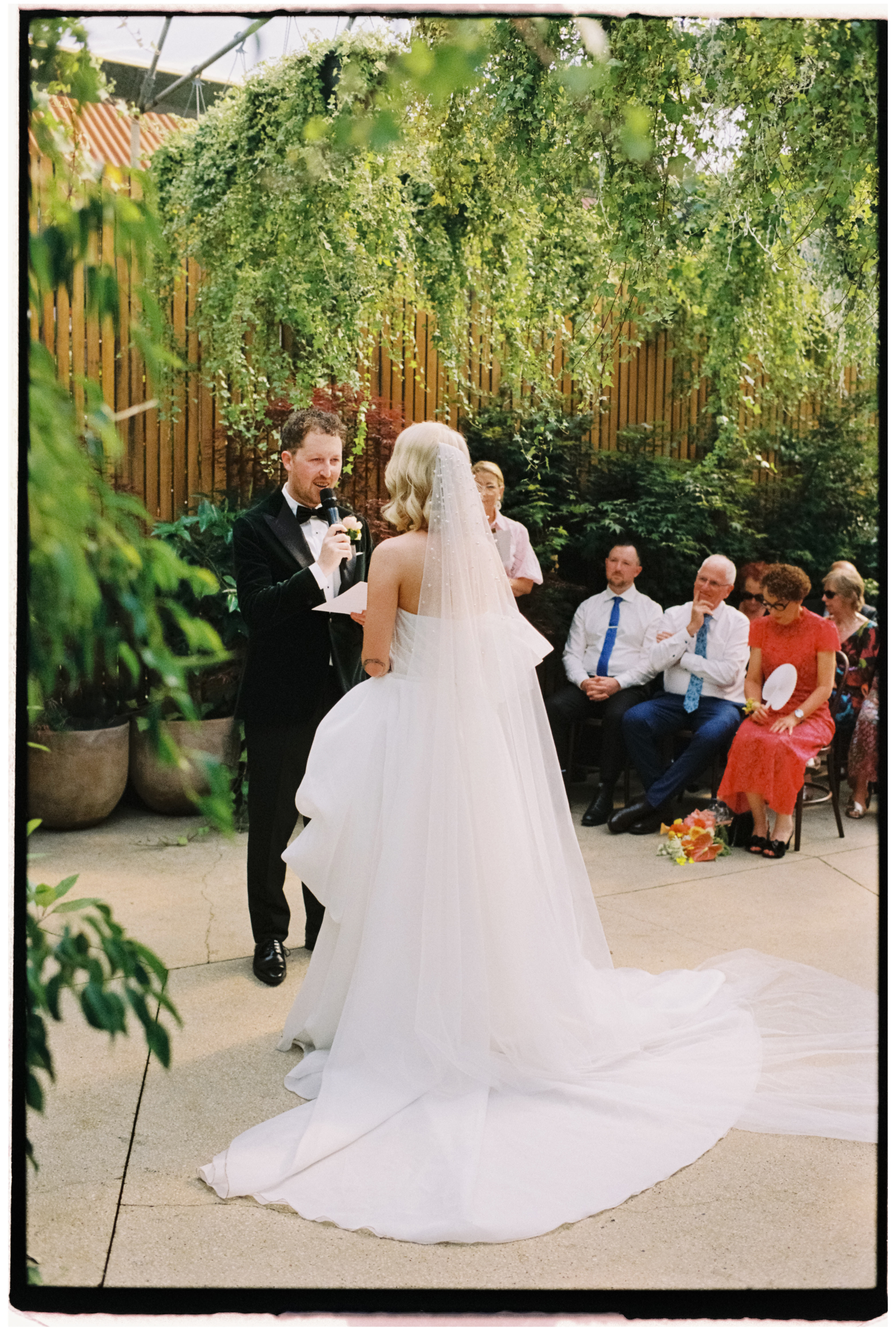 The groom reads his personal vows to the bride while his family watches on at Glasshaus Outside in Richmond Melbourne.