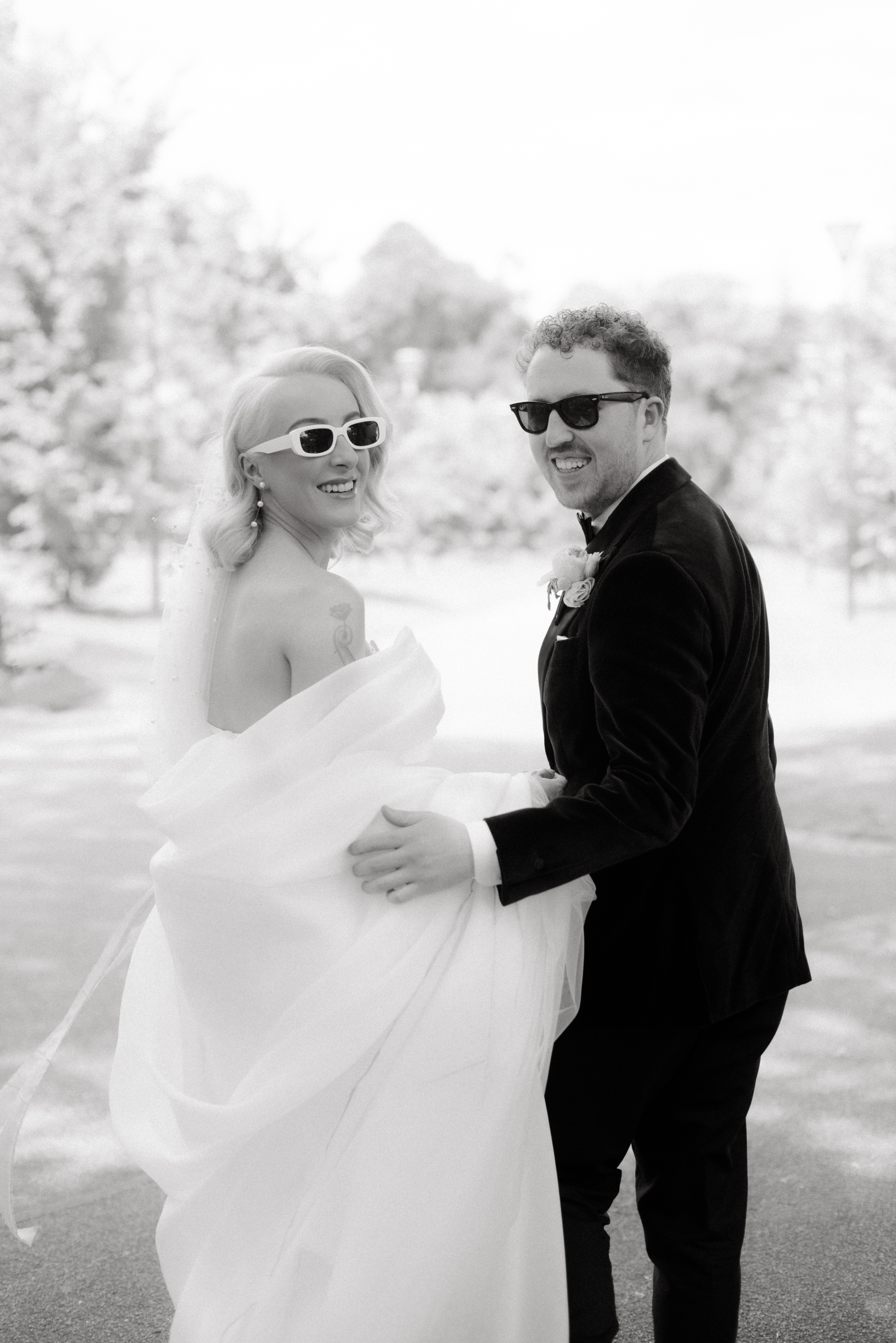 A black and white photo of a bride and groom wearing sunglasses walking through Fitzroy Gardens during their photo portrait session.