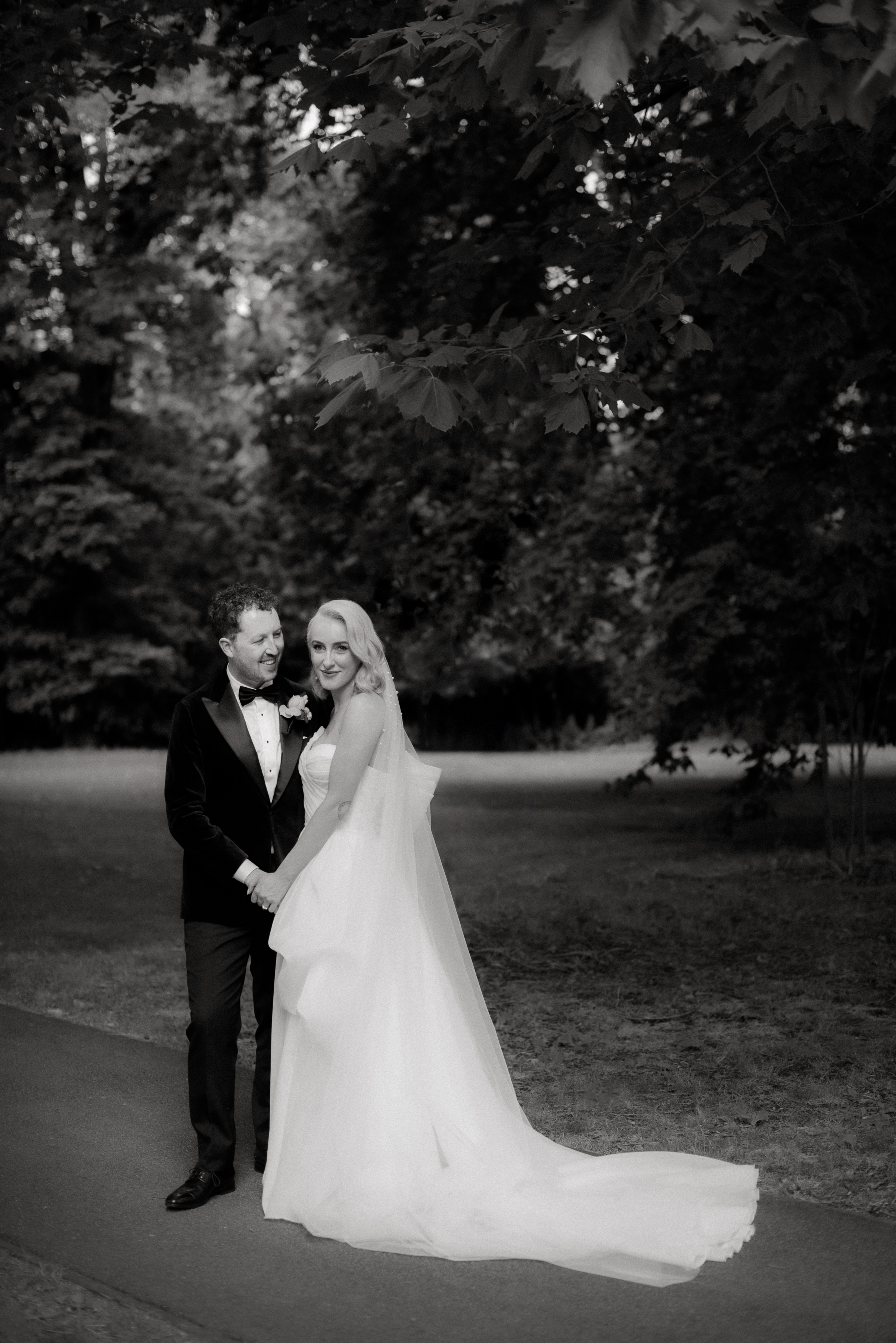 A black and white photo of a bride and groom in front of a row of trees at Fitzroy Gardens during their photo portrait session.