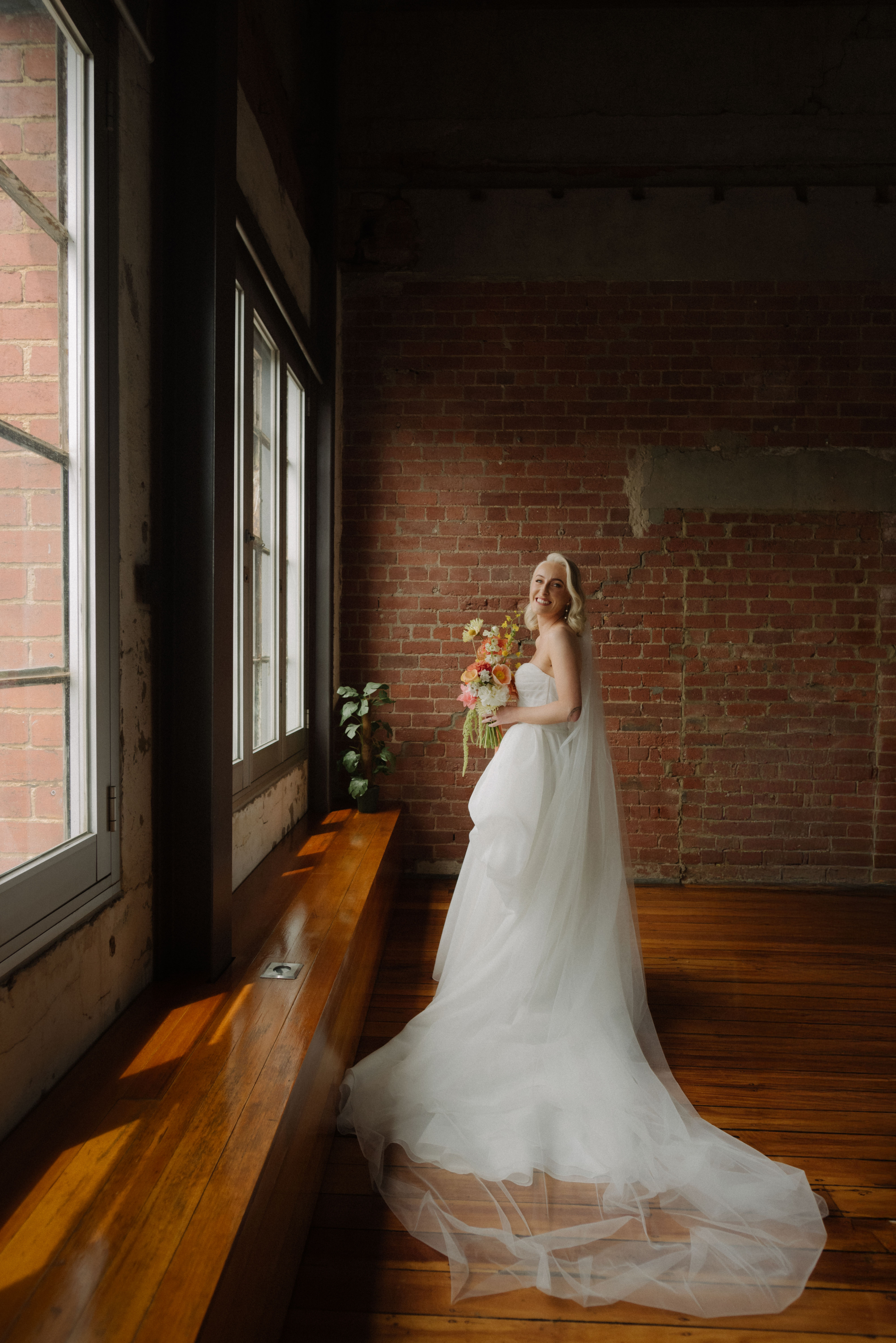 A bride smiles in front of a red brick wall, her white bridal dress and veil drapes elegantly along the floor.